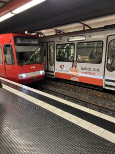 This picture shows a picture of two trams. One is leaving the station heading towards Bad Godesburg. Another tram colored red with the number 16 on it is pulling into the station. This one is headed towards Cologne.