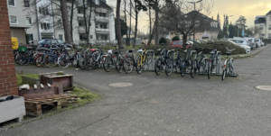 This is a picture of a bike rack completely filled with bikes around sunset by some houses and the train station. 