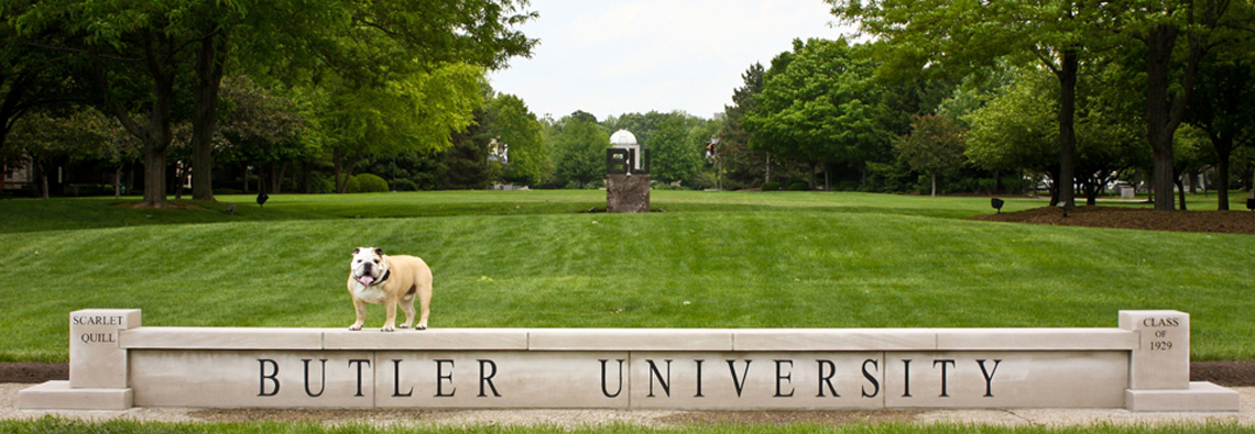 The Butler bulldog stands atop the Butler University sign, with a large grass field in the background.