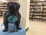 A sculpture of a bulldog with a blue-and-green rendition of the earth on its chest. Library stacks containing DVDs can be seen in the background.