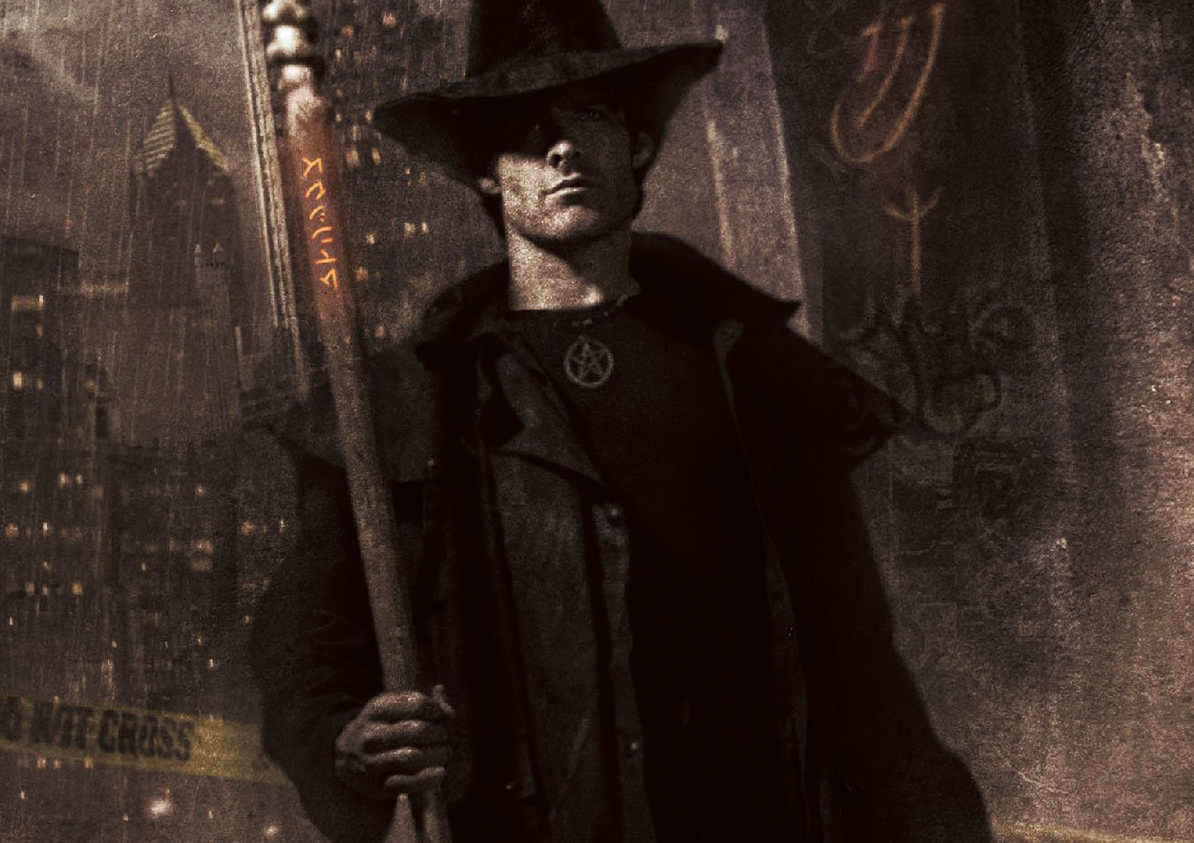 A male human wearing a black overcoat, black hat, and a wizard staff stands in front of a grey/brown urban background.